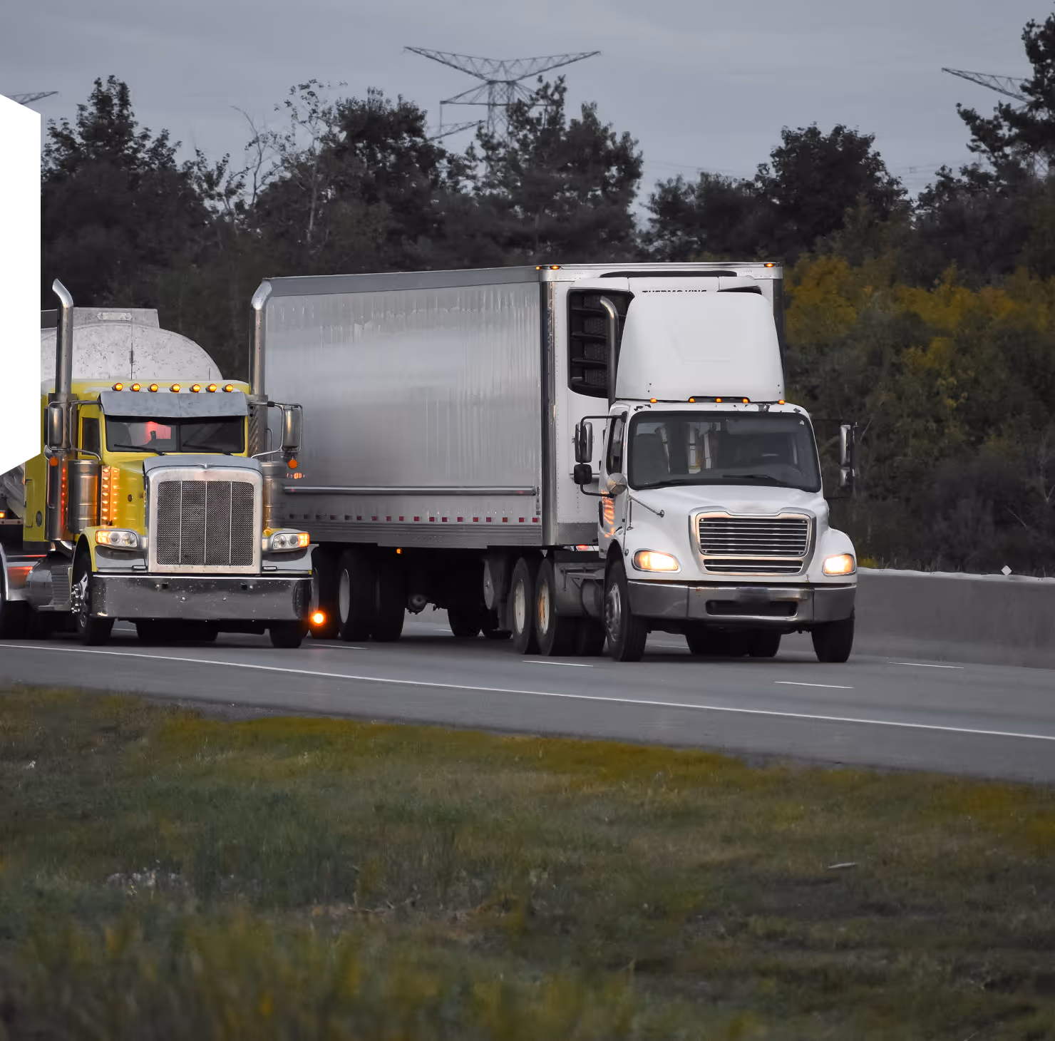 Two semi-trailer trucks driving side by side on a highway with trees and power lines in the background.